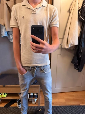 Beige pikétröja från Polo Ralph Lauren - Säljer en klassisk beige pikétröja från Polo Ralph Lauren med korta ärmar och den ikoniska loggan broderad på bröstet. Tröjan har krage och knappar vid halsen. Perfekt till jeans eller shorts för en clean och stilren look.
