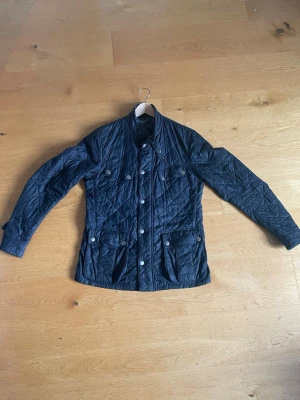 Marinblå quiltad jacka med knappar - Snygg marinblå quiltad jacka med knappar framtill och flera fickor. Jackan har lång ärm och en klassisk krage. Perfekt för dig som vill ha en stilren och tidlös look. Materialet är lätt och passar bra till både vår och höst. Hänget har lossnat som man kan se på bild 4 och jackan har en skada på höger framficka (bild 5).