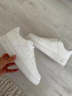 Vita klassiska Nike Air Force 1 - Säljer ett par helvita Nike Air Force 1 sneakers i lågt utförande. Skorna har en stilren design med perforerad tå, vita skosnören och diskret Nike-logga på sidan. Tillverkade i slätt läder med gummisula för grym komfort och streetstyle.