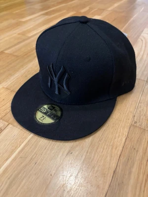 Svart New Era Yankees keps 59FIFTY - Snygg svart keps från New Era, modell 59FIFTY, med New York Yankees-logga broderad framtill i svart. Klassisk flat brim och strukturerad passform. Tillverkad i slitstarkt tyg som ger en clean och stilren look.