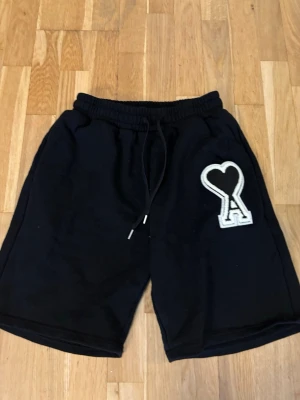 Svarta shorts från Ami med hjärtdetalj - Svarta shorts från Ami med elastisk midja och snörning. På vänster ben finns en stor vit hjärtdetalj med bokstaven A. Sköna och avslappnade, perfekta för sommaren. Tillverkade i mjuk bomull.