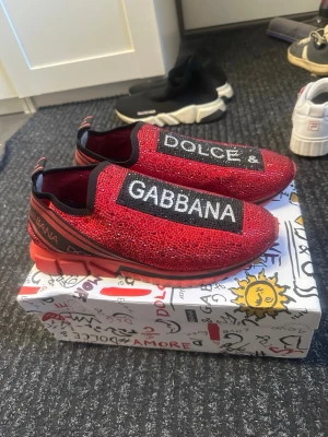 Röda Dolce & Gabbana sneakers - Snygga röda sneakers från Dolce & Gabbana med glittrande stenar över hela ovandelen och tydliga logotexter på ovansidan. Skorna har svarta och vita detaljer samt elastisk slip-on design. Sulan är chunky och röd, vilket ger en cool och modern look. bra skick smutsiga och inte använda på länge, snöret där de står dolce gabbana på har gått av på båda sidorna där bak men det är inget som spelar någon roll egentligen. finns med på bilderna.
