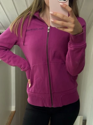 Rosa hoodie från Peak Performance - Lila/rosa hoodie från Peak Performance med dragkedja framtill och broderad logga på bröstet. 
