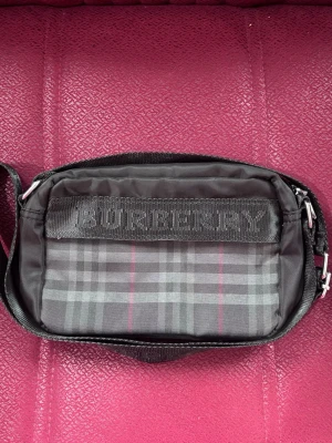 Burberry maglväska i rutigt tyg - Säljer en snygg axelväska från Burberry i klassiskt rutigt mönster i grått, svart och vinrött. Väskan har justerbar axelrem med Burberry-logga och detaljer i svart tyg. Perfekt storlek för det viktigaste och riktigt trendig design. Kan bara mötas! Stockholm!