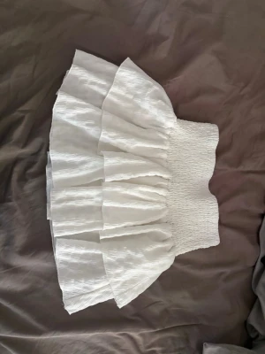 Vit volangkjol med smock - Supersöt vit kjol med bred smock i midjan och flera lager volanger. Kjolen är kort och har en luftig känsla, perfekt för varma dagar. Materialet ser ut att vara lätt och något strukturerat för extra volym.