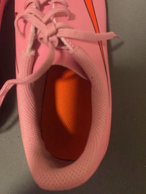 Rosa fotbollsskor med snörning - Snygga fotbollsskor i rosa med orange innersula och svarta detaljer. Skorna har klassisk snörning och en låg profil som ger bra passform. Materialet är syntetiskt och designen är sportig och modern, perfekt för dig som vill sticka ut på planen.