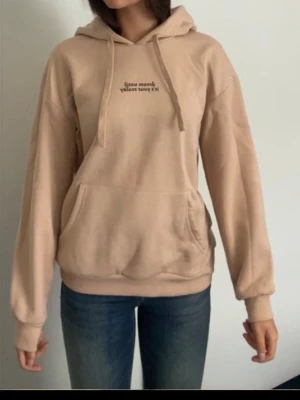 Beige hoodie med tryck och huva - Snygg beige hoodie med huva och dragsnören. Framtill finns ett litet tryck med text och en stor känguruficka. Hoodien har ribbade muddar vid ärmslut och nederkant. Perfekt för en chill och avslappnad stil.
