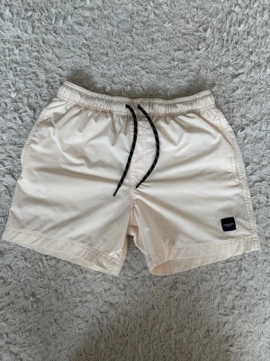 Beige badshorts från Jack & Jones - Snygga beige badshorts från Jack & Jones med svart dragsko i midjan och liten svart logotyp-patch på vänster ben. Klassisk modell med elastisk midja och sidfickor. Perfekta för stranden eller poolen.