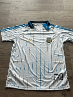 Argentina Adidas fotbollströja blåvit - Snygg Argentina fotbollströja från Adidas med klassiska blå ränder på vit botten, blå detaljer på axlar och krage samt broderad landslagssköld. Tillverkad i 100% polyester, perfekt för match eller träning.