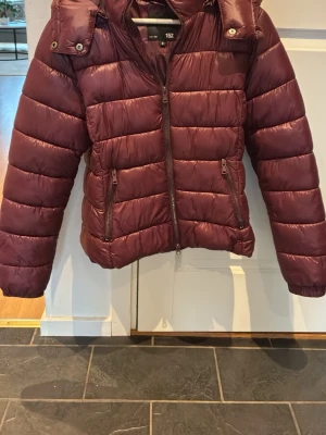 Vinröd pufferjacka med huva - Snygg vinröd pufferjacka med avtagbar huva och två dragkedjeförsedda fickor framtill. Jackan har en quiltad design och stängs med dragkedja. Perfekt för kalla vinterdagar. Skön, fin och bra jacka 