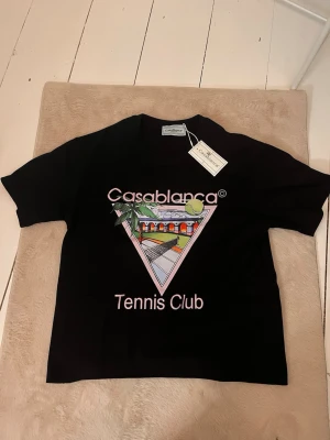 Svart Casablanca Tennis Club t-shirt - Svart t-shirt från Casablanca med färgglatt tryck framtill som visar en tennisbana och texten 'Tennis Club'. T-shirten har rund hals och korta ärmar. Perfekt för dig som gillar sportig och trendig stil.