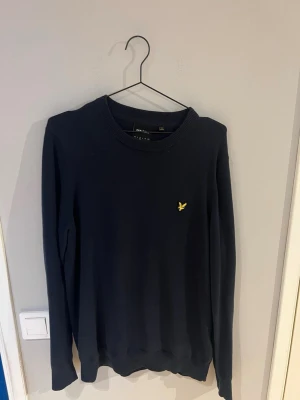 Marinblå tröja från Lyle & Scott - Snygg marinblå tröja från Lyle & Scott med klassisk gul logga på bröstet. Tröjan har rund halsringning och långa ärmar. Perfekt för dig som gillar stilrena och tidlösa plagg. Materialet känns mjukt och bekvämt.