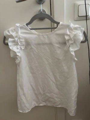 Vit blus med volangärmar - Söt vit blus med volangdetaljer på ärmarna. Blusen har rund halsringning och är tillverkad i ett lätt tyg som ger en luftig känsla. Perfekt för dig som gillar en romantisk och trendig stil.