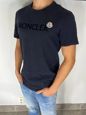 Moncler t-shirt  - Snygg svart t-shirt från Moncler med stor logga och text på bröstet. Klassisk rund halsringning och korta ärmar. Tillverkad i mjuk bomull som känns skön mot huden. Perfekt till jeans eller shorts för en clean och stilren look.