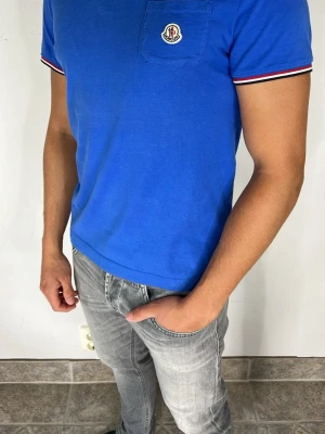 Blå Moncler t-shirt med bröstficka - Snygg blå t-shirt från Moncler med klassisk bröstficka och logga. T-shirten har korta ärmar med randiga detaljer i rött, vitt och svart vid ärmsluten. Tillverkad i mjuk bomull och har en normal passform. Perfekt till jeans eller shorts.