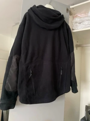 Svart hoodie i tjock fleece med dragkedja och huva  - Svart hoodie i fleece-material med dragkedja och stor huva. Har förstärkta partier på armbågarna och två fickor med dragkedja på sidorna. Perfekt för dig som gillar en enkel men funktionell stil.