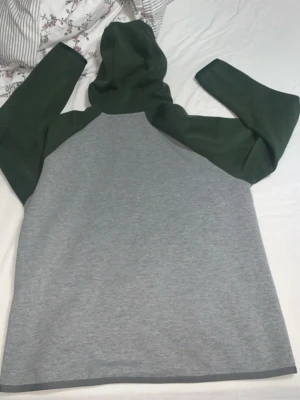 nike tech fleece - Nike hoodie i grått med mörkgröna ärmar och huva. Dragkedja framtill och klassisk Nike-logga på bröstet. Tillverkad i mjukt bomullsmaterial, perfekt för en avslappnad streetstyle-look.