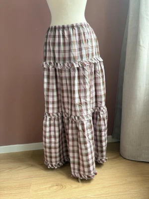 Rutig långkjol med volangdetaljer - Långkjol stl S  i rutigt mönster med volanger och resår i midjan. Kjolen har en luftig passform och är sydd i ett lätt material med rosa, beige och gröna toner. Perfekt för dig som gillar en romantisk och avslappnad stil.