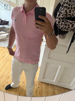 Ljusrosa pikétröja ralph lauren - Snygg ljusrosa pikétröja med klassisk krage och korta ärmar. Tröjan har en blå broderad logga på bröstet och är tillverkad i mjukt bomullsmaterial. Perfekt för en stilren och fräsch look.tröjan är i storlek L och är i jättefint skick helt utan defekter och slitage!