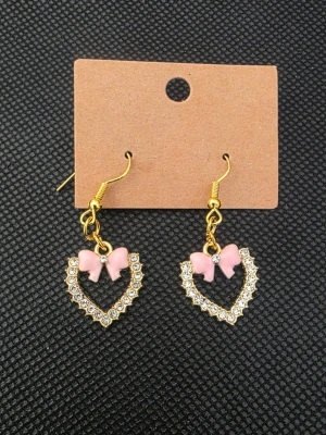 Hjärformade örhängen med rosa rosett - Snygga örhängen i guldton med hjärtformad design, dekorerade med glittrande stenar och en söt rosa rosett upptill. Perfekta för dig som gillar gulliga accessoarer med lite extra bling.