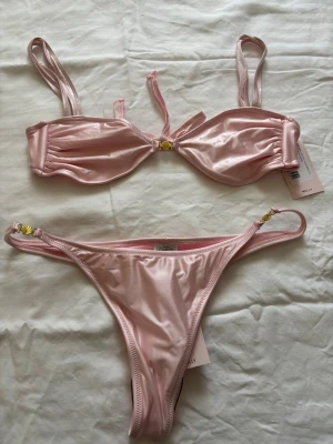 Ljusrosa bikini med gulddetaljer - Helt oanvänd, inte ens testat på. Snygg ljusrosa bikini i glansigt material med guldfärgade detaljer på både överdel och underdel. Överdelen har smala axelband och en liten rosett framtill, medan underdelen har hög skärning och rynk bak. Perfekt för sommaren och strandhäng!