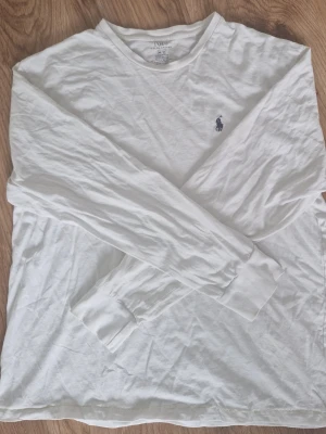 Ralph Lauren tröja - Klassisk vit långärmad tröja från Polo Ralph Lauren med rund halsringning och den ikoniska mörkblå logotypen broderad på bröstet. Tillverkad i mjuk bomull som känns skön mot huden. Perfekt till jeans eller under en jacka.