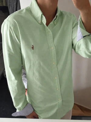 Grön skjorta från Ralph Lauren - Snygg ljusgrön skjorta från Ralph Lauren i slim fit-modell. Skjortan har klassisk krage med knappar, långa ärmar och den ikoniska broderade loggan på bröstet. Tillverkad i bomull och har randigt tyg på insidan av kragen och manschetterna.