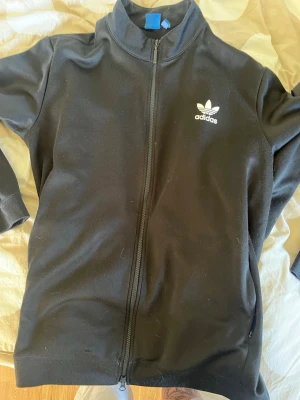 Svart Adidas ziptröja med tryck - Snygg svart ziptröja från Adidas med klassisk vit logga på bröstet och texten 'THE BRAND WITH THE 3 STRIPES' i vitt på ryggen. Tröjan har hög krage, hel dragkedja och två sidofickor. Perfekt till streetwear-stilen.