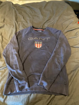 Mörkblå sweatshirt från GANT - Mörkblå sweatshirt från GANT med broderad logga och amerikanskinspirerat sköldmärke på bröstet. Tröjan har rund halsringning och ribbade muddar vid ärmslut och nederkant. Perfekt för en avslappnad och stilren look.