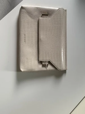 Beige kuvertväska i krokopräglat skinn - Stilren beige kuvertväska i krokopräglat konstläder med unik asymmetrisk lockdesign och guldfärgat spänne. Väskan har en elegant och modern look som passar perfekt till många olika outfits.