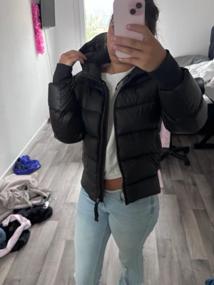 Svart vinterjacka - Säljer en svart pufferjacka med huva och dragkedja framtill. Jackan har en klassisk quiltad design och breda kanaler som ger en riktigt puffig look. Perfekt för kalla dagar och enkel att matcha med olika outfits.