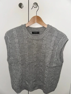 Grå stickad kabelväst - Säljer en grå stickad väst med klassiskt kabelstickat mönster och ribbade kanter. Perfekt att slänga över en skjorta eller t-shirt för en chill och trendig look. Rund halsringning och loose fit för skön vibe.
