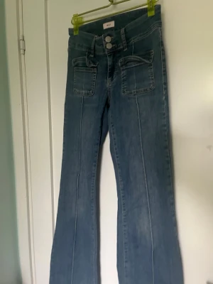 Blå bootcut jeans från Nelly - Snygga blå bootcut jeans från Nelly med två stora fickor framtill och dubbla knappar i midjan. Jeansen har en klassisk denimlook och markerade sömmar längs benen. Perfekta för dig som gillar en retro vibe och vill sticka ut lite extra.