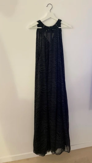 Svart långklänning med zebra-mönster - Snygg svart långklänning med zebramönster och nitar vid halsringningen. Klänningen har ärmlös design och ett luftigt, svepande tyg som ger en cool look. Perfekt för dig som vill sticka ut med en unik stil.