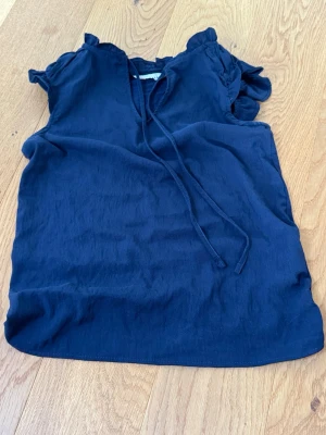 Mörkblå blus från Mennaf Paris - Supersöt mörkblå blus från Mennaf Paris med volangdetaljer vid ärmslut och knytband i halsen. Blusen är ärmlös och har en avslappnad passform, perfekt till jeans eller kjol. Materialet känns lätt och luftigt.