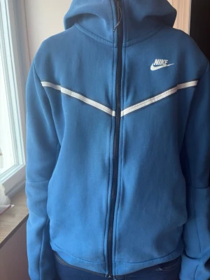 Blå Nike hoodie med dragkedja - Snygg blå hoodie från Nike med vit logga på bröstet och vita ränder över bröstet. Dragkedja framtill och huva med snörning. Ficka med dragkedja på ena ärmen för extra stilpoäng. Perfekt för en sportig och avslappnad look.