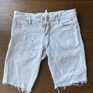 Vita jeansshorts från Dsquared2 - Säljer ett par vita jeansshorts från Dsquared2 med slitna detaljer och råa kanter. Shortsen har klassisk femficksdesign, knappgylf och en röd Dsquared2-logga vid gylfen. Perfekta för en avslappnad och trendig look. W28, skinny/slim fit