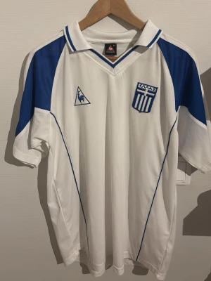 Vit och blå Grekland fotbollströja - Säljer en vit fotbollströja från Le Coq Sportif med blå detaljer och Greklands landslagsemblem på bröstet. Tröjan har korta ärmar, v-ringad krage med blå ränder och är tillverkad i ett lätt och ventilerande material. Perfekt för match eller träning. Denna är samma tema som den andra tröjan jag har lagt upp fast en vit borta tröja. 