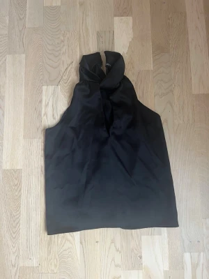 Svart ärmlös sidenblus med knyt - Stilren svart blus i siden med hög krage och knyt i halsen. Ärmlös modell som ger en elegant och modern look. Perfekt att matcha med både jeans och kjol för en trendig outfit.
