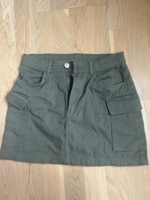 Grön kortkjol med fickor från H&M - Snygg grön kortkjol från H&M med stora sidfickor och bälteshällor. Kjolen har en rak passform och stängs med dragkedja och knapp framtill. Perfekt för en avslappnad och trendig look.