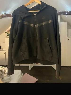 Svart Nike zip hoodie med huva - Säljer en svart zip hoodie från Nike med två svarta ränder över bröstet och diskret Nike-logga på vänster sida. Hoodien har dragkedja framtill, huva och två stora fickor. Perfekt för en sportig och avslappnad stil.