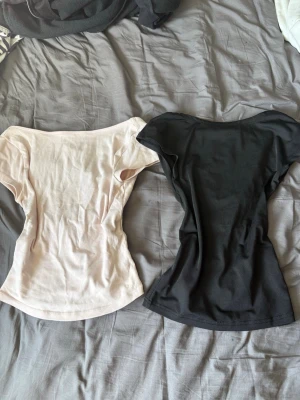 Två bas-toppar i svart och beige - Säljer två enkla bas-toppar i svart och beige. Båda har rund halsringning och korta ärmar. Perfekta att matcha med jeans eller kjol. Materialet känns mjukt och stretchigt, troligtvis bomull eller bomullsblandning.