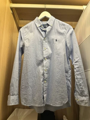 Ljusblå skjorta från Polo Ralph Lauren - Klassisk ljusblå skjorta från Polo Ralph Lauren i slim fit-modell. Skjortan har långa ärmar, knappar framtill och den ikoniska broderade loggan på bröstet. Perfekt för en stilren och clean look. Storlek 16 years old.