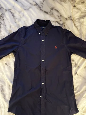 Mörkblå skjorta från Ralph Lauren - Klassisk mörkblå skjorta från Ralph Lauren i slim fit-modell. Skjortan har långa ärmar, knappar framtill och en röd broderad logga på bröstet. Tillverkad i bomull och passar perfekt till en stilren look.