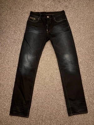 G-star jeans - Säljer ett par svarta jeans med raka ben och snygga slitningar på knän och bakfickor. Jeansen har klassisk femficksdesign, kontrastsömmar och en mörk tvätt med lätt blekta partier. Perfekta för en avslappnad och trendig stil. Storlek 32:34