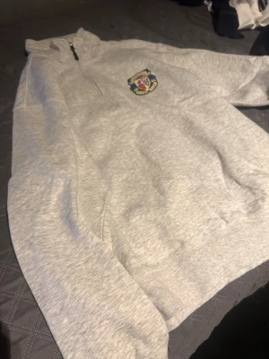 Grå halvzip hoodie Djurgår'n - Grå hoodie med halv dragkedja och broderad Djurgår'n-logga på bröstet. Mjuk bomullskänsla och klassisk passform. Perfekt för dig som vill visa ditt lagstöd med stil.