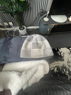 Gråvit rutmönstrad mössa - Säljer en stilren gråvit mössa med rutmönster och diskret textdetalj framtill. Mössan är stickad och har en mjuk, bekväm känsla som passar perfekt för kyliga dagar. Enkel att matcha med olika outfits och ger en trendig touch till din look.