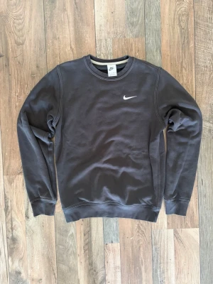 Svart sweatshirt från Nike - Klassisk svart sweatshirt från Nike med rund halsringning och vit broderad Swoosh-logga på bröstet. Tröjan har ribbade muddar vid ärmslut och nederkant. Perfekt för en avslappnad och sportig stil.