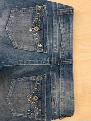 Blå utsvängda Lågmidjade jeans med pärldetaljer - Säljer ett par blå jeans med utsvängda ben och snygga slitningar. Fickorna är dekorerade med pärlor, nitar och små stjärnor för en unik look. midja: ca 40 cm. Innerben: ca 81 cm