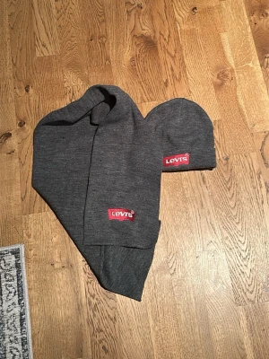 Mörkgrå halsduk från Levi's och mössa  - Säljer en stilren mörkgrå halsduk o mössa från Levi's med klassisk röd logotyp. Båda är ribbstickad och mjuk, perfekt för kalla dagar. Passar dig som vill ha en enkel men snygg accessoar till vintern.